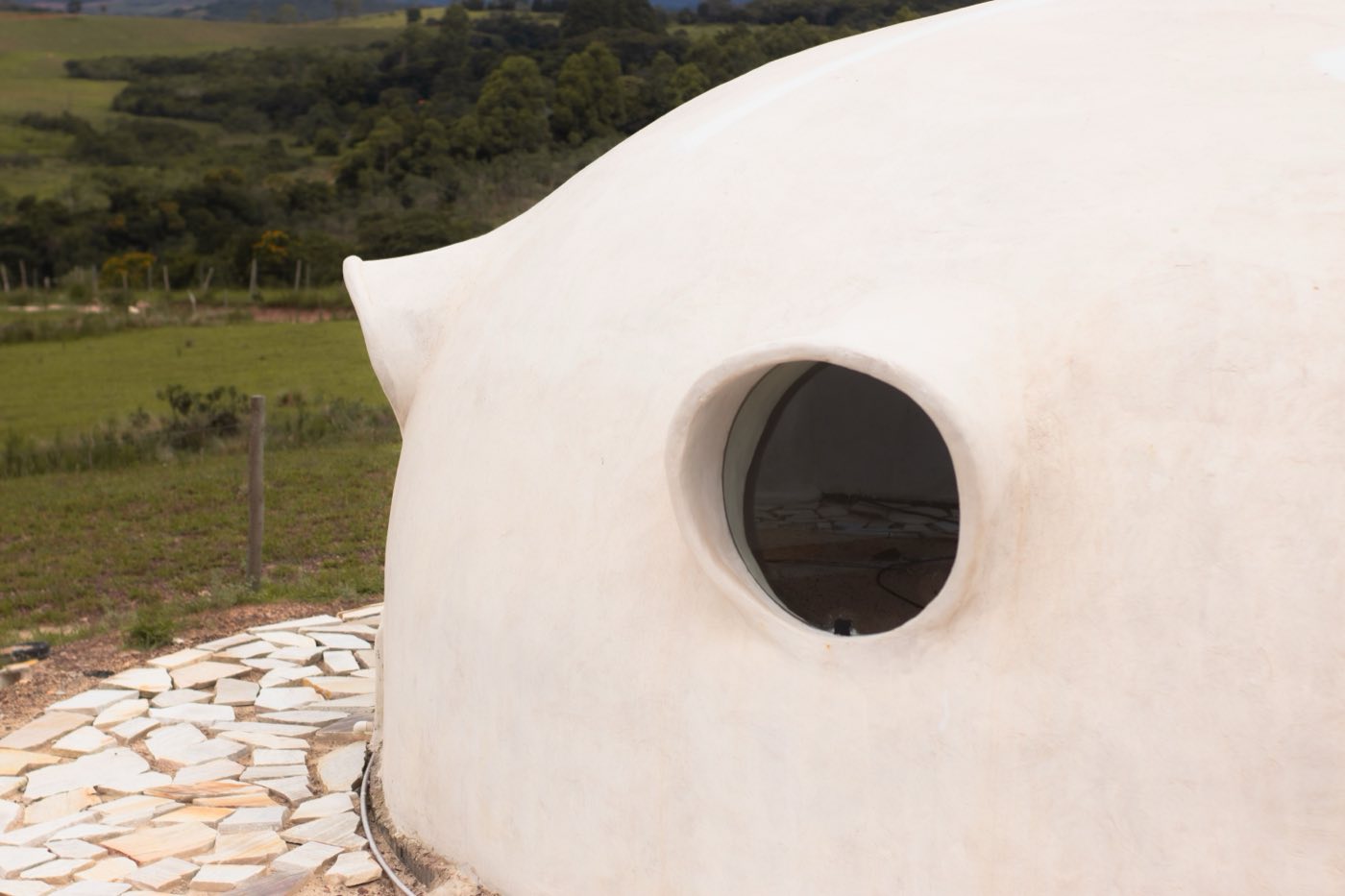 A curved, plastered earthen wall with a round window opening set on a stone base
