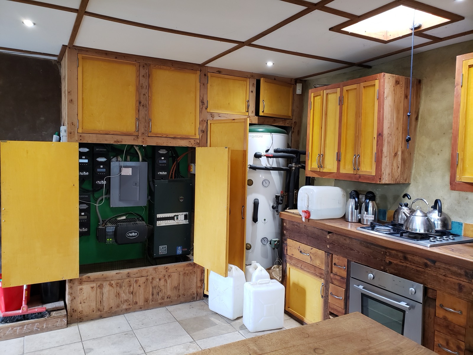 Earthship kitchen with cabinet doors open, revealing charge controllers, an inverter and a breaker panel beside a hot water cylinder