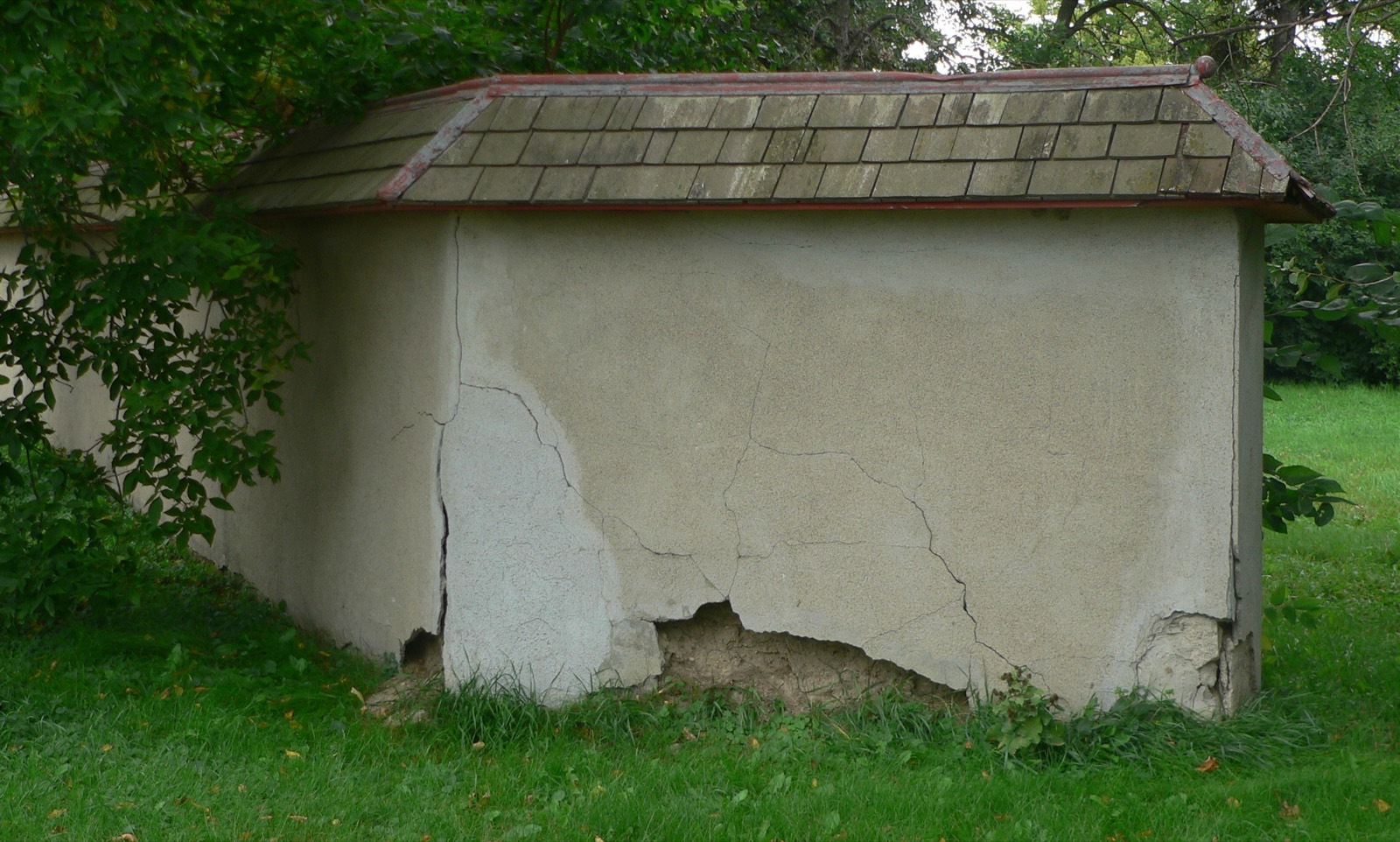 A rammed earth outbuilding with a deep tiled roof overhang, its cement render cracked and broken away along the base