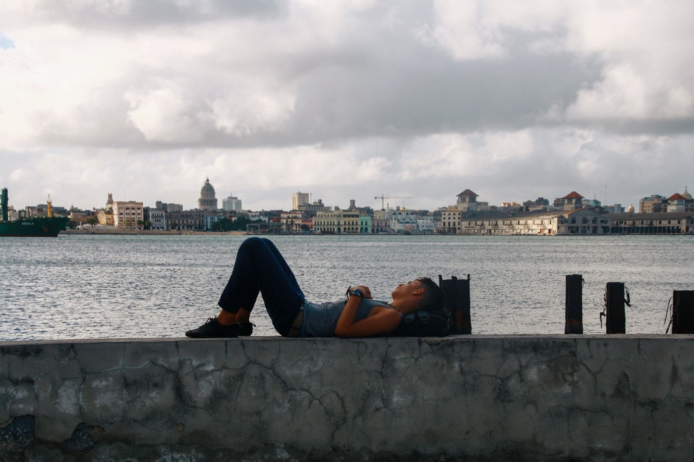 A person lying on the Havana malecon wall with the city skyline and Capitolio dome across the harbour